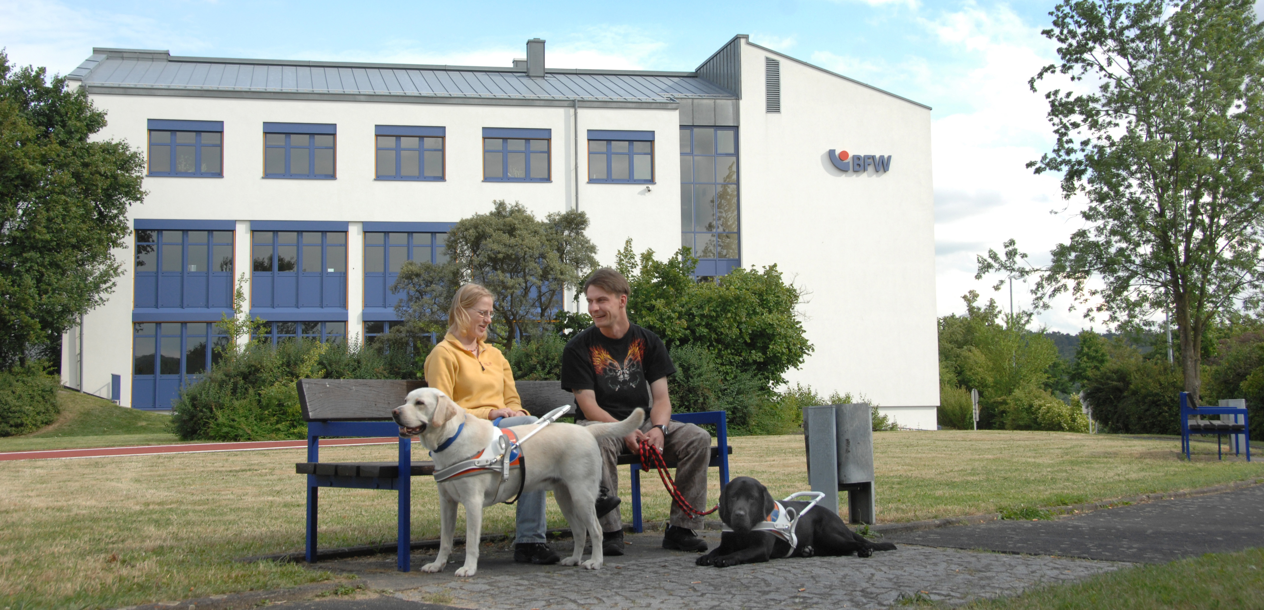 Foto zwei blinde Teilnehmer*innen mit Führhunden auf einer Bank auf dem Gelände